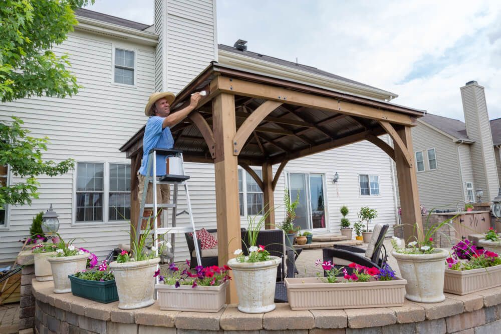 Man staining a backyard gazebo on a ladder viewed over flowers on a patio wall looking towards a large suburban house