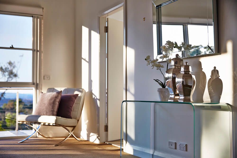 bedroom corner with a glass console table with decorative objects, a mirror, white leather Barcelona chair with cushions
