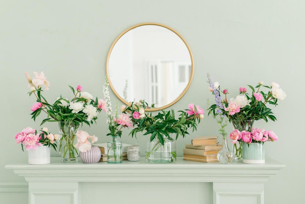 Pink and white peonies on the bookshelf in glass vases. Round golden mirror above the fireplace