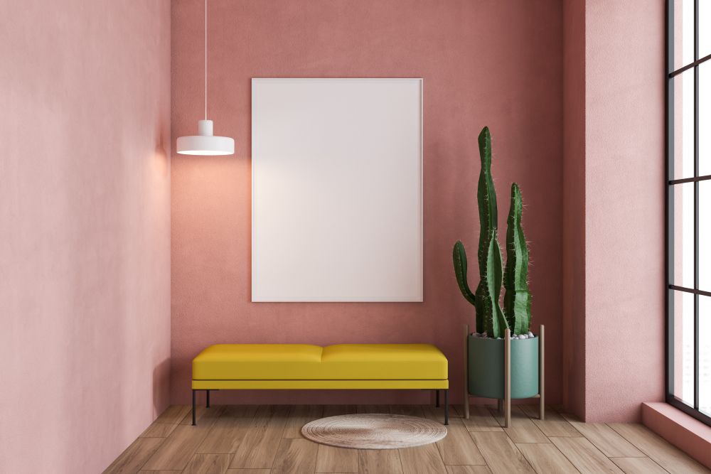ein weißes Bild, ein gelber Stuhl, ein kleiner runder Teppich und ein Kaktus an einer rosafarbenen Wand