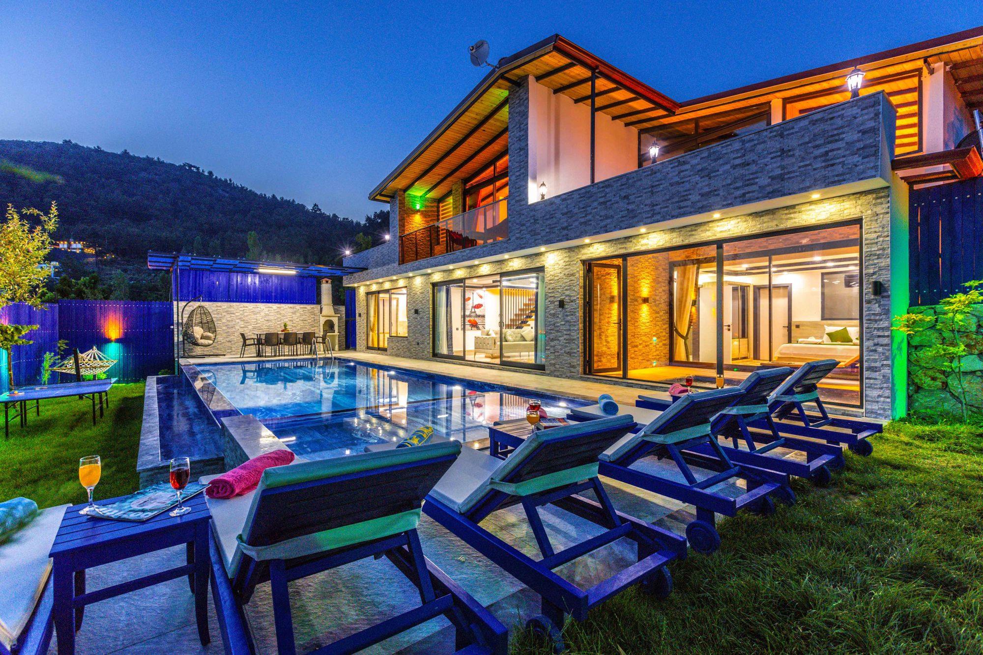 Vista noturna de uma casa contemporânea com piscina iluminada e mobiliário exterior flexível, evidenciando um estilo de vida luxuoso e sofisticado. 