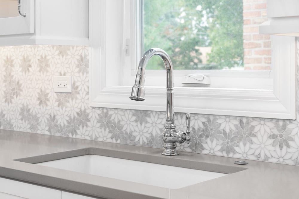 A kitchen faucet with a marble daisy flower tiled backsplash, white cabinets, chrome faucet, and a light brown quartz countertop.
