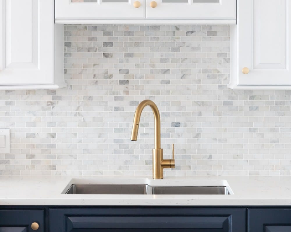 white and blue kitchen with a gold faucet, marble countertop, and small marble subway tile backsplash