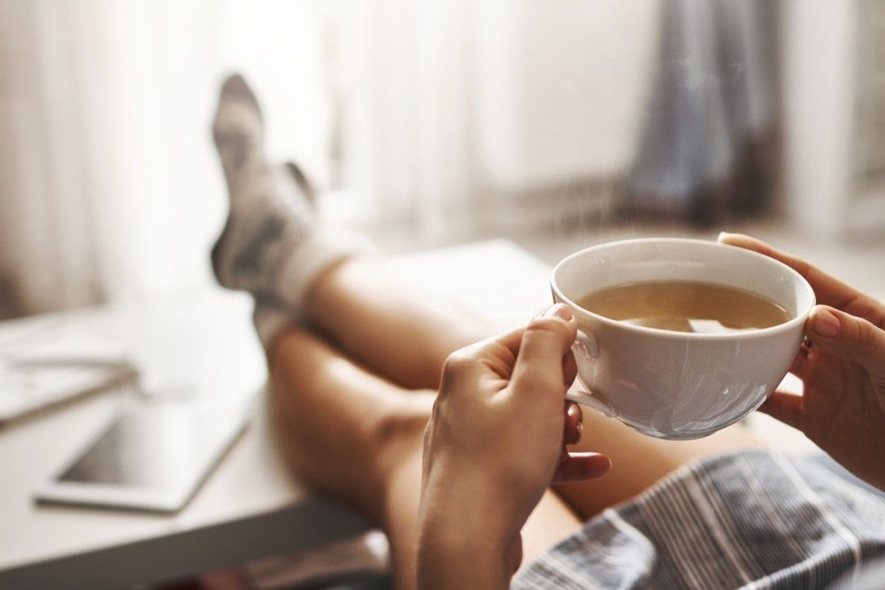 Woman lying on couch, feet on coffee table, drinking hot tea and enjoying sunlight