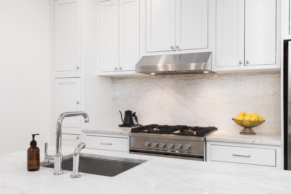 A kitchen detail with white cabinets, stainless steel stove and hood, marble countertops and backsplash,
