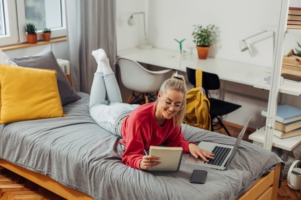 student on a bed in a dorm with a desk behind her