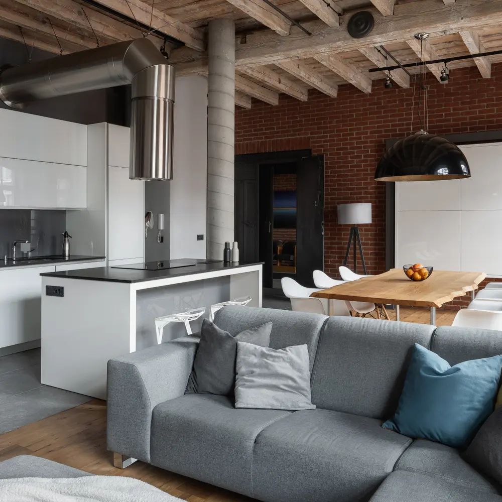 Mieszkanie w stylu industrialnym z szarą narożną sofą, stołem jadalnym i otwartą kuchnią.