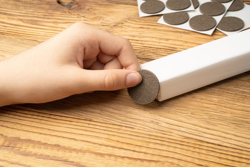 Holzmöbel-Pflege: Die Hand eines Mädchens, das ein selbstklebendes Filzkissen auf das Bein eines Holzstuhls klebt.