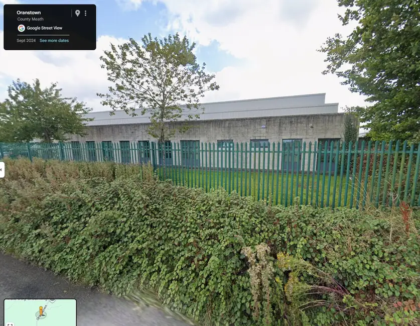 Neighboring building behind a green metal fence, with trees and vegetation in the foreground.