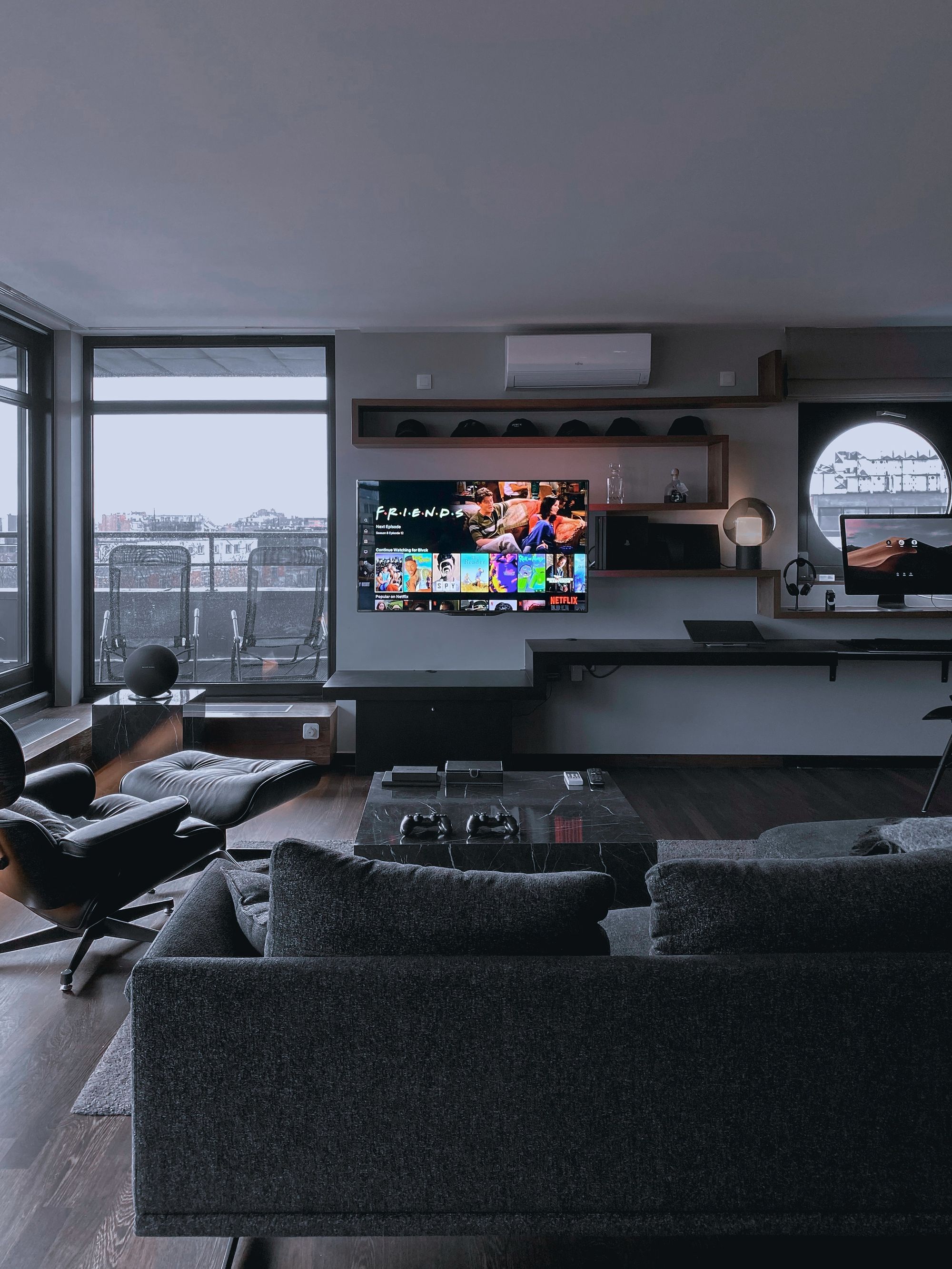 un salon avec un canapé gris, une table basse en marbre noir et un fauteuil, face à un écran diffusant Friends 