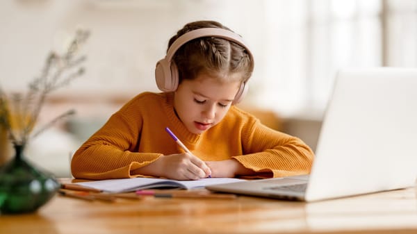 kleines Mädchen, das seine Hausaufgaben neben einem Laptop macht und Kopfhörer trägt